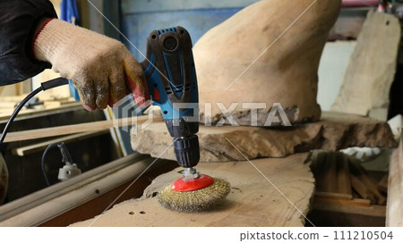 carpenter's hand holds a screwdriver with a metal round attachment for sanding wooden material, processing wood for production in the workshop using a sanding metal brush on a drill carpenter's hand holds a screwdriver with a metal round attachment for sanding wooden material, processing wood for production in the workshop using a sanding metal brush on a drill 111210504