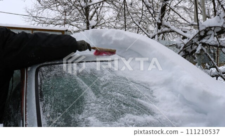 clearing snow from a car with a brush, a hand clears a thick layer of snow from the roof and windshield of a gray car, the consequences of a snow storm and snowfall for transport 111210537