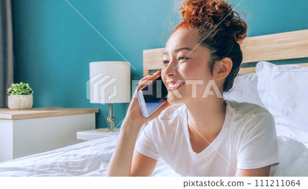 beautiful Asian woman with red curly hair in casual tee talking on the phone in bed, 16:9 widescreen image 111211064
