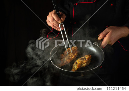 The cook fries carp fish in a frying pan in a restaurant kitchen. Idea of a delicious fish diet on a black background. Fork in the chef hand The cook fries carp fish in a frying pan in a restaurant kitchen. Idea of a delicious fish diet on a black background. Fork in the chef hand 111211464