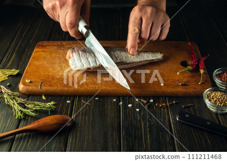 Hands of a chef using a knife to cut fresh Hake fish on a wooden cutting board. Home-cooked national fish dish according to a unique recipe Hands of a chef using a knife to cut fresh Hake fish on a wooden cutting board. Home-cooked national fish dish according to a unique recipe 111211468
