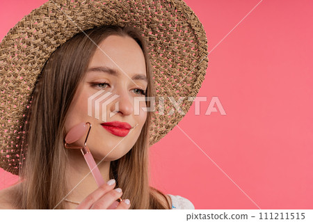 Young woman with rose quartz stone roller on pink background. Facial self care 111211515