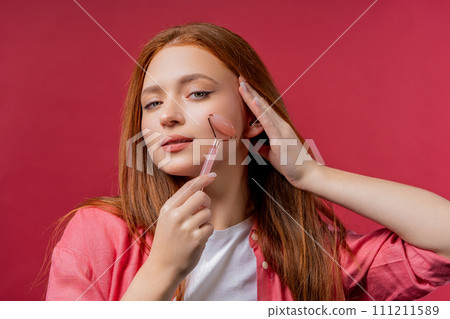 Young woman with rose quartz stone roller on pink background. Facial self care 111211589