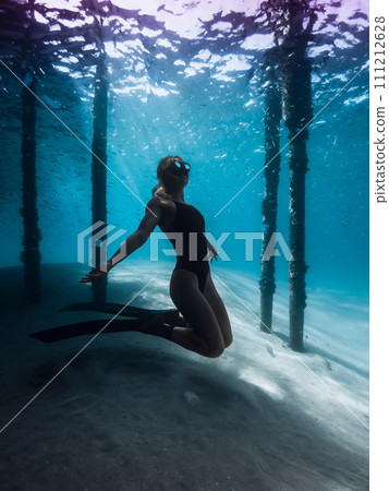 Freediver woman posing underwater under the pier in blue ocean. Female swims with fins under the pier 111212628