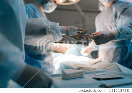 Closeup hands of unrecognizable nurse assists surgeons, giving surgical clamp with tampon during surgical operation. Doctors team working performing surgery in intensive care unit 111213718