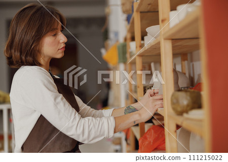 Pretty female ceramist standing near shelves of clay mugs while working in pottery studio 111215022