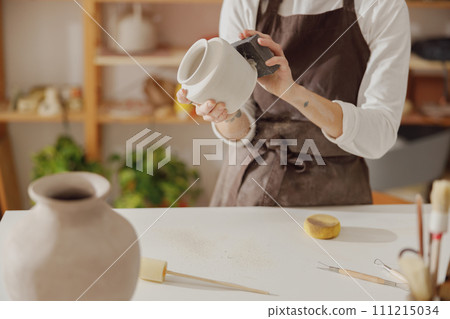 Close up of female ceramist wearing apron work with unfired clay vase in pottery studio Close up of female ceramist wearing apron work with unfired clay vase in pottery studio 111215034
