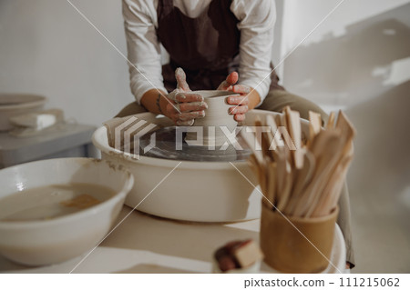 Female potter in apron making shape of clay vase on spinning pottery tool in ceramic workshop Female potter in apron making shape of clay vase on spinning pottery tool in ceramic workshop 111215062
