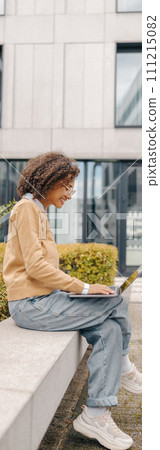 Smiling woman manager in eyeglasses is working on laptop sitting on background of office building Smiling woman manager in eyeglasses is working on laptop sitting on background of office building 111215082
