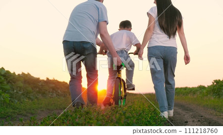 Father grasps bicycle seat providing support to kid pedaling by mother on plantation 111218760