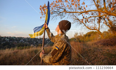 Young man in military uniform waving flag of Ukraine against beautiful sunset at background. Male ukrainian army soldier lifted national banner at countryside. Victory against russian aggression 111230664