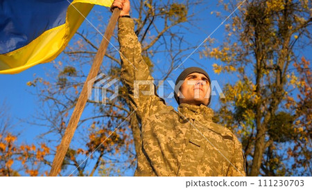 Male ukrainian army soldier lifting blue-yellow banner in honor of the victory against russian aggression. Young man in camouflage uniform raising a waving flag of Ukraine at countryside. End of war 111230703