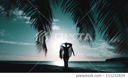 Woman walking on night tropical beach. Silhouette of slim girl under the palm coconut tree raise hands up. Bright sky with sun rays reflected in water. Blue filter toning. Travel, tourism Woman walking on night tropical beach. Silhouette of slim girl under the palm coconut tree raise hands up. Bright sky with sun rays reflected in water. Blue filter toning. Travel, tourism 111232834