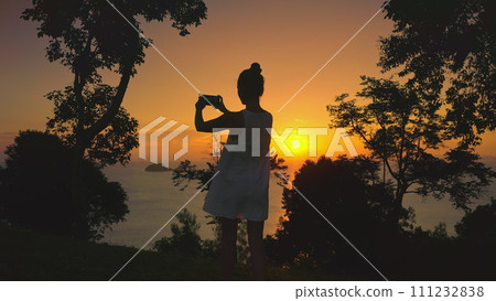 Woman make sunset panorama enjoy sea sunset view. Woman silhouette standing in forest take photo on mobile phone. Bright orange sky sun in background. Exotic nature landscape. Travel, holiday relax 111232838