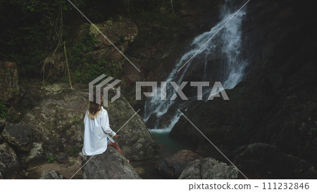 Female relax on stone looking jungle waterfall. Woman in white shirt enjoy beautiful wild nature landscape, exotic rainforest. Outdoor lifestyle travel summer holiday vacation. Dark toning, drone shot Female relax on stone looking jungle waterfall. Woman in white shirt enjoy beautiful wild nature landscape, exotic rainforest. Outdoor lifestyle travel summer holiday vacation. Dark toning, drone shot 111232846