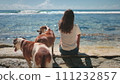 Woman with two brown dogs relax sitting on sea coast beach. Hugging dogs on the beach and looking at the ocean surf waves together. People and pets enjoying weekend time, quiet relax, beautiful nature 111232857