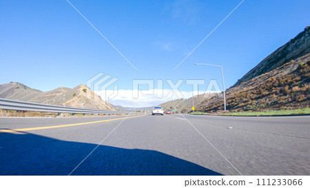 On a crisp winter day, a car cruises along the iconic Highway 1 near San Luis Obispo, California. The surrounding landscape is brownish and subdued, with rolling hills and patches of coastal 111233066