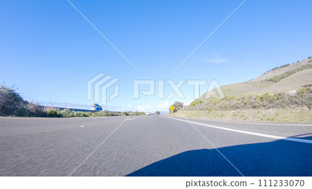 On a crisp winter day, a car cruises along the iconic Highway 1 near San Luis Obispo, California. The surrounding landscape is brownish and subdued, with rolling hills and patches of coastal 111233070