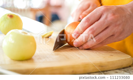 In the welcoming setting of a modern kitchen, a young man continues his dinner preparation process. He's currently involved in slicing yellow onions into rings, prepping them for grilling on an In the welcoming setting of a modern kitchen, a young man continues his dinner preparation process. He's currently involved in slicing yellow onions into rings, prepping them for grilling on an 111233080