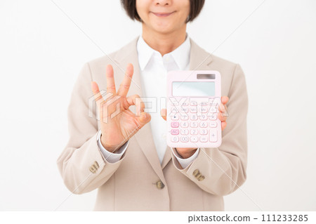 A middle-aged business woman wearing a beige suit holding a pink calculator and making a circle sign with her finger 111233285