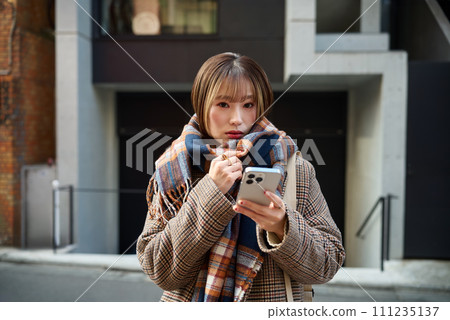 A young woman in winter clothes wearing a scarf holding a smartphone 111235137