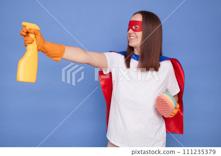 Smiling brown haired woman in superhero costume mask and protective rubber gloves posing isolated over blue background holding sponge and spray raised arm with detergent advertising cleaning service 111235379