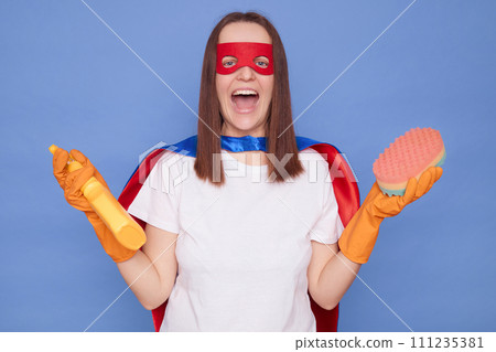 Crazy overjoyed woman wearing superhero costume mask and protective rubber gloves posing isolated over blue background holding detergent and sponge exclaiming enjoying her work in cleaning service 111235381