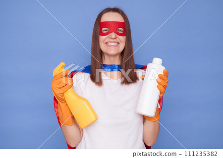 Cheerful brown haired woman wearing superhero costume mask and protective rubber gloves posing isolated over blue background holding sanitary supplies being ready to help with household chores 111235382