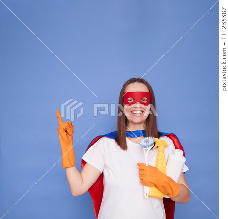 Smiling cheerful satisfied adult woman cleaner wearing gloves and superhero costume holding detergent isolated over blue background pointing away at copy space for advertisement 111235387