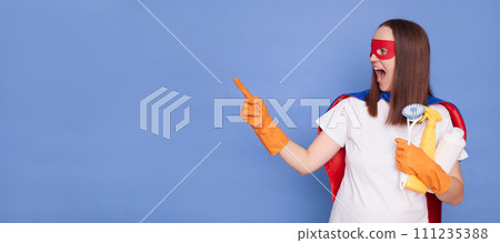 Overjoyed amazed excited adult woman cleaner wearing gloves and superhero costume holding sanitary spray presenting advertisement of cleaning service isolated over blue background Overjoyed amazed excited adult woman cleaner wearing gloves and superhero costume holding sanitary spray presenting advertisement of cleaning service isolated over blue background 111235388
