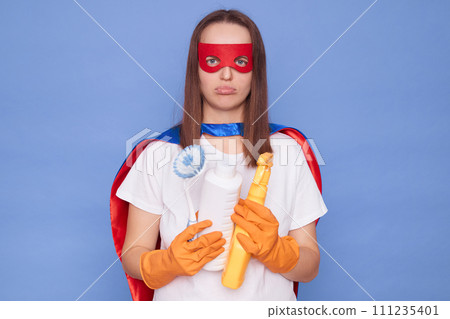 Exhausted adult woman cleaner wearing gloves and superhero costume holding cleaning spray being tired of doing household chores isolated over blue background Exhausted adult woman cleaner wearing gloves and superhero costume holding cleaning spray being tired of doing household chores isolated over blue background 111235401