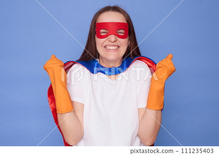 Overjoyed Caucasian brown haired woman wearing superhero costume white T-shirt and rubber gloves clenched fists rejoicing her success isolated over blue background Overjoyed Caucasian brown haired woman wearing superhero costume white T-shirt and rubber gloves clenched fists rejoicing her success isolated over blue background 111235403