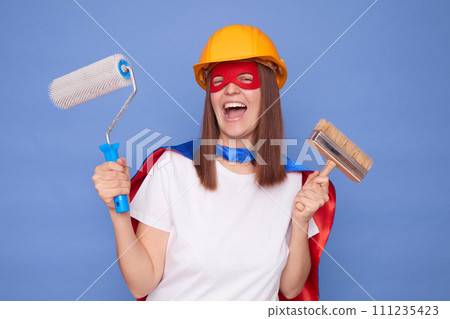 Professional painter. Decorative painting. Building service. Overjoyed woman painter wearing superhero costume and protective helmet holding painting roller and brush isolated over blue background Professional painter. Decorative painting. Building service. Overjoyed woman painter wearing superhero costume and protective helmet holding painting roller and brush isolated over blue background 111235423