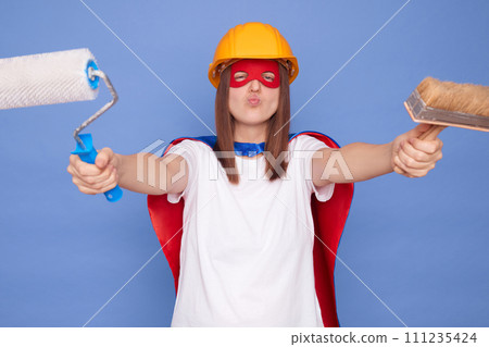 Funny woman painter wearing superhero costume and protective helmet holding painting roller and brush isolated over blue background working sending air kiss with pout lips 111235424