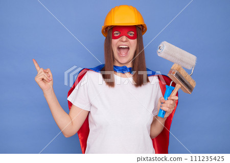 Renovating home. Paintbrush technique. Amazed woman painter wearing superhero costume and protective helmet holding painting roller and brush isolated over blue background pointing finger away 111235425