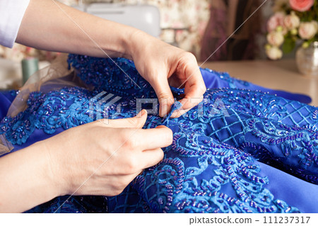 Seamstress at work. Dressmaker making clothes in modern studio. Tailor holding pencil and marking fabric. Woman standing at table with cut textile, sewing machine, thread, pins, needles, tape, cutouts 111237317