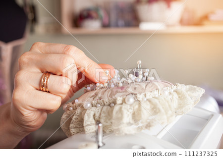 Seamstress at work. Dressmaker making clothes in modern studio. Tailor holding pencil and marking fabric. Woman standing at table with cut textile, sewing machine, thread, pins, needles, tape, cutouts Seamstress at work. Dressmaker making clothes in modern studio. Tailor holding pencil and marking fabric. Woman standing at table with cut textile, sewing machine, thread, pins, needles, tape, cutouts 111237325