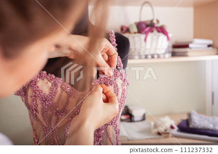 Seamstress at work. Dressmaker making clothes in modern studio. Tailor holding pencil and marking fabric. Woman standing at table with cut textile, sewing machine, thread, pins, needles, tape, cutouts Seamstress at work. Dressmaker making clothes in modern studio. Tailor holding pencil and marking fabric. Woman standing at table with cut textile, sewing machine, thread, pins, needles, tape, cutouts 111237332