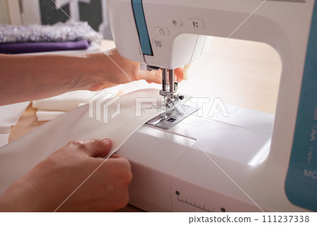Seamstress at work. Dressmaker making clothes in modern studio. Tailor holding pencil and marking fabric. Woman standing at table with cut textile, sewing machine, thread, pins, needles, tape, cutouts 111237338