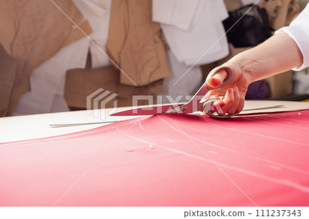 Seamstress at work. Dressmaker making clothes in modern studio. Tailor holding pencil and marking fabric. Woman standing at table with cut textile, sewing machine, thread, pins, needles, tape, cutouts Seamstress at work. Dressmaker making clothes in modern studio. Tailor holding pencil and marking fabric. Woman standing at table with cut textile, sewing machine, thread, pins, needles, tape, cutouts 111237343