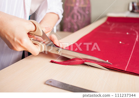Seamstress at work. Dressmaker making clothes in modern studio. Tailor holding pencil and marking fabric. Woman standing at table with cut textile, sewing machine, thread, pins, needles, tape, cutouts Seamstress at work. Dressmaker making clothes in modern studio. Tailor holding pencil and marking fabric. Woman standing at table with cut textile, sewing machine, thread, pins, needles, tape, cutouts 111237344