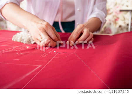 Seamstress at work. Dressmaker making clothes in modern studio. Tailor holding pencil and marking fabric. Woman standing at table with cut textile, sewing machine, thread, pins, needles, tape, cutouts Seamstress at work. Dressmaker making clothes in modern studio. Tailor holding pencil and marking fabric. Woman standing at table with cut textile, sewing machine, thread, pins, needles, tape, cutouts 111237345