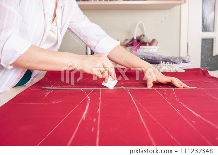 Seamstress at work. Dressmaker making clothes in modern studio. Tailor holding pencil and marking fabric. Woman standing at table with cut textile, sewing machine, thread, pins, needles, tape, cutouts 111237348