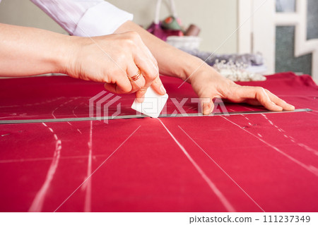 Seamstress at work. Dressmaker making clothes in modern studio. Tailor holding pencil and marking fabric. Woman standing at table with cut textile, sewing machine, thread, pins, needles, tape, cutouts 111237349