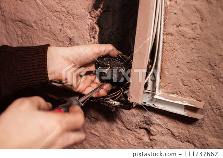 A man fixes a burnt socket. Short circuit, burnt wires. traces of smoke fire on the wall. The electrical outlet was completely melted, the voltage drop from the use of an electric heater. Screwdriver A man fixes a burnt socket. Short circuit, burnt wires. traces of smoke fire on the wall. The electrical outlet was completely melted, the voltage drop from the use of an electric heater. Screwdriver 111237667