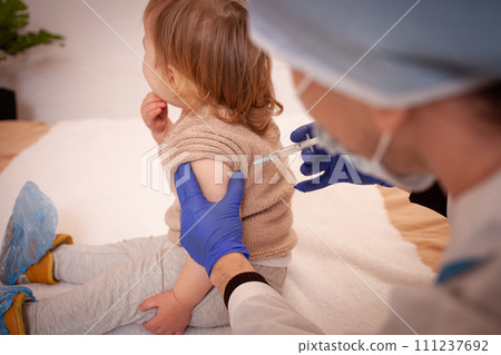 The doctor vaccinates the child against coronavirus. The child is crying and afraid. A man in a robe, hat, mask and gloves makes an injection to the child. Home quarantine 111237692