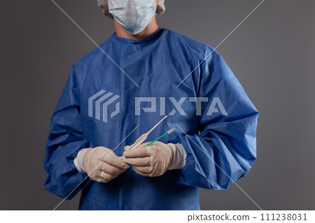 A gynecologist with a venus mirror in his hands. Gynecological accessories, brush, stick. A doctor in a medical mask, a robe, gloves, with a gynecological mirror on a gray background. A gynecologist with a venus mirror in his hands. Gynecological accessories, brush, stick. A doctor in a medical mask, a robe, gloves, with a gynecological mirror on a gray background. 111238031