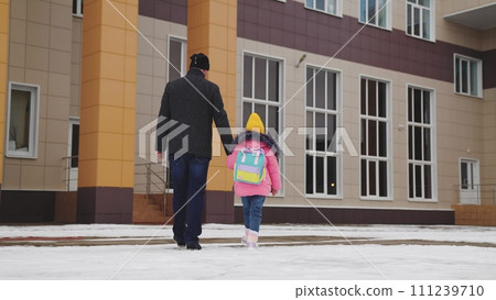 father takes child school holding his hand. happy family. snowy road school. father leads his little girl with school backpack through snow school. cold season. Dad holds hand child kid with backpack. 111239710