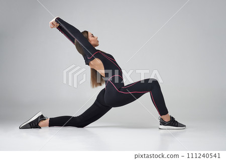 Flexible female in black tracksuit performing Warrior pose of yoga against grey background. Side view of young Caucasian woman practicing yoga asana on floor, isolated. Concept of yoga, lifestyle. 111240541