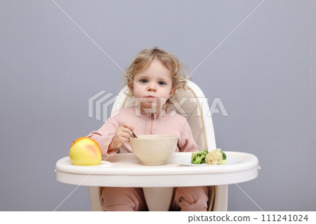 Funny little baby girl eating healthy food while sitting in high chair chewing soup looking at camera with curios facial expression isolated over gray background Funny little baby girl eating healthy food while sitting in high chair chewing soup looking at camera with curios facial expression isolated over gray background 111241024